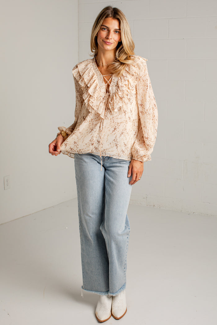 A woman modeling a light beige blouse with ruffles and a tie neckline, paired with light blue wide-leg jeans and white ankle boots, standing in a bright white studio.