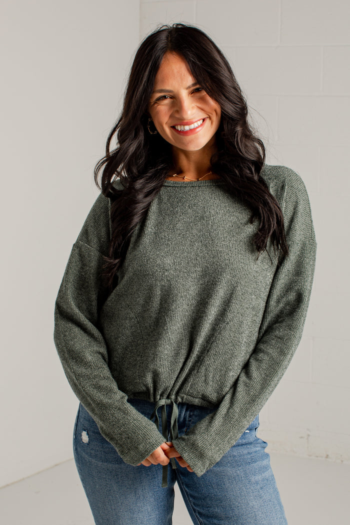 A woman modeling a loose, heathered sage green knit sweater with a tied hem, paired with blue jeans and brown heeled sandals, standing against a white studio wall.