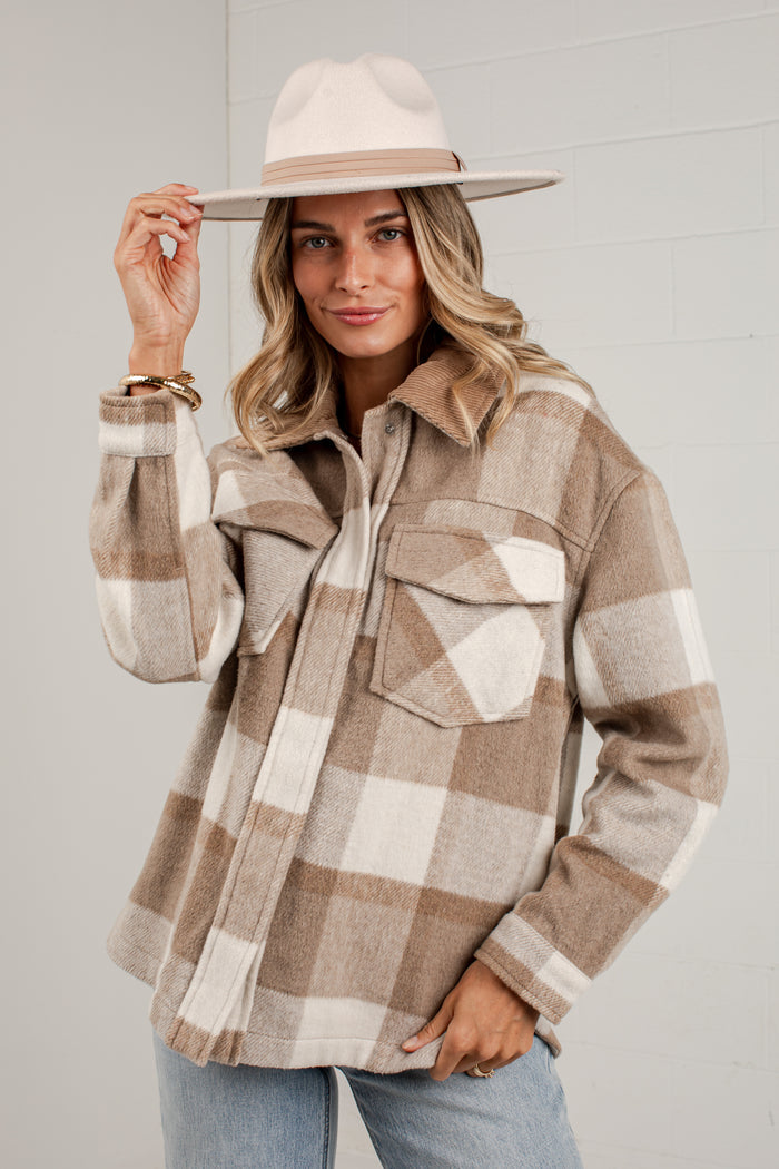 A woman wearing a beige and brown plaid flannel button-up shirt jacket with a wide-brimmed cream hat, posing against a light gray brick wall.