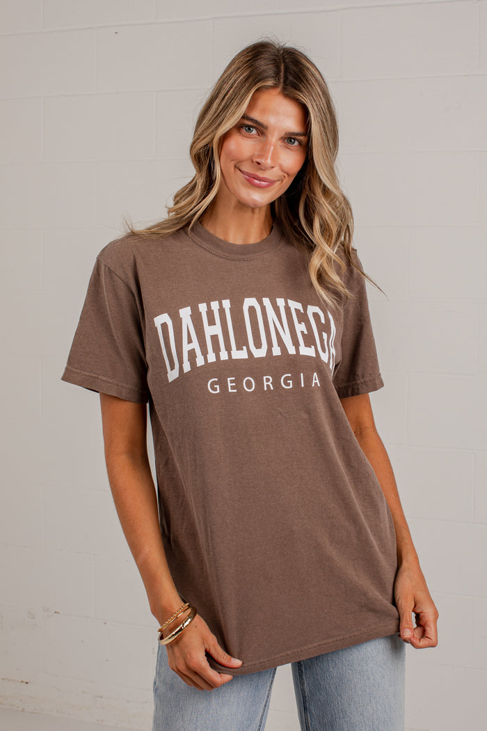 A woman wearing a brown short-sleeve graphic t-shirt with 'DAHLONEGA GEORGIA' printed in white across the chest, paired with light blue jeans, standing against a white brick wall.