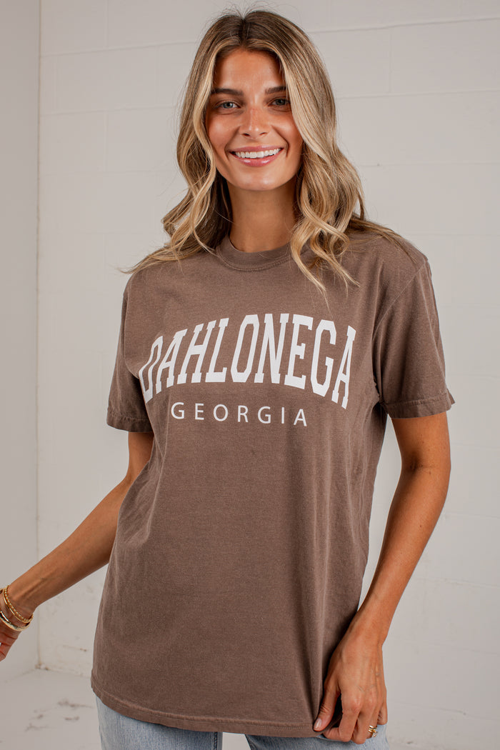 A woman wearing a brown short-sleeve graphic t-shirt with 'DAHLONEGA GEORGIA' printed in white arched letters across the chest, posed against a white paneled wall.