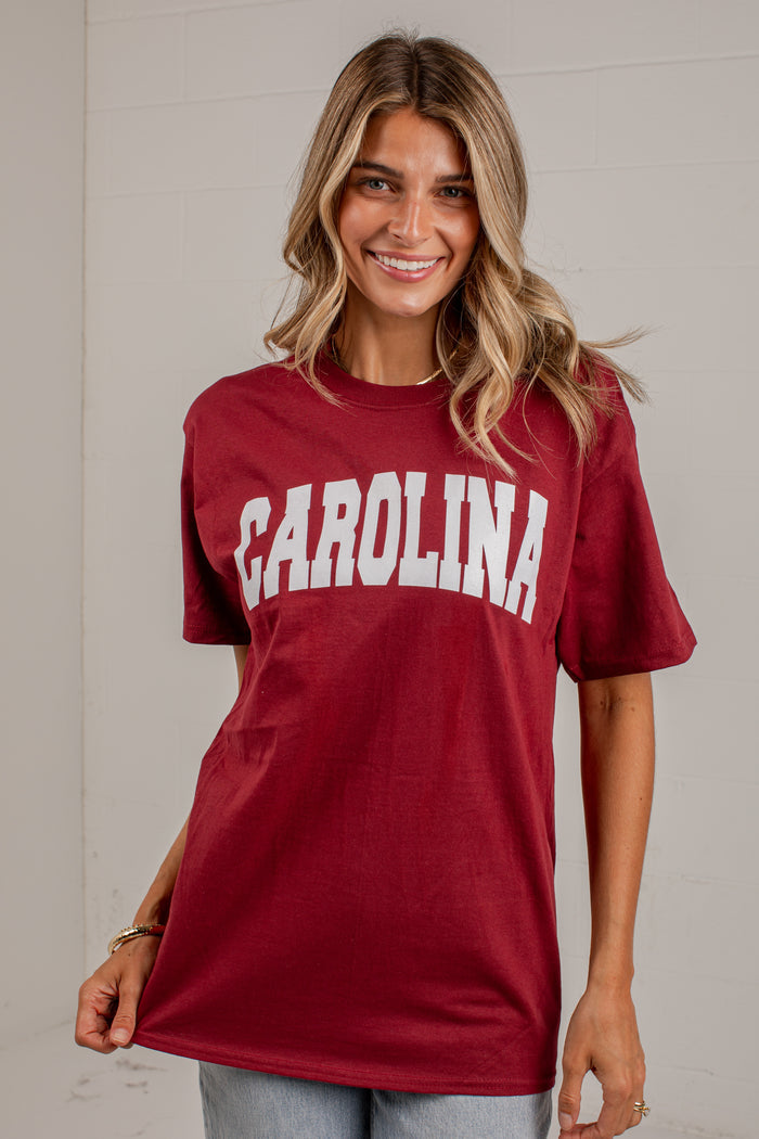 A woman wearing a maroon short-sleeve t-shirt with 'CAROLINA' printed in white arched letters across the chest, paired with light blue jeans, standing against a light gray paneled wall.