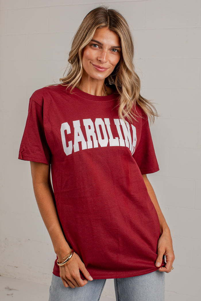 A woman wearing a maroon short-sleeve t-shirt with 'CAROLINA' printed in white arched block letters across the chest, paired with light blue jeans, set against a light gray brick wall.