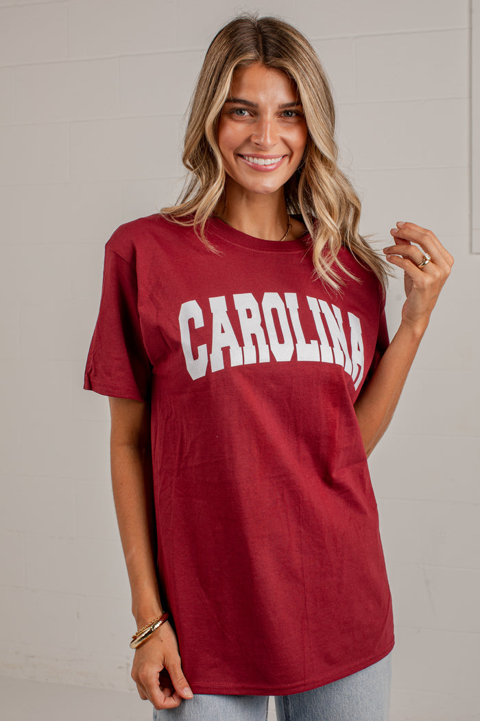 A woman wearing a maroon short-sleeve t-shirt with 'CAROLINA' printed in white arched block letters across the chest, paired with light blue jeans, standing against a light gray brick wall.