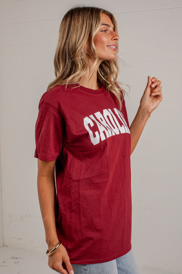 A woman wearing a maroon short-sleeve graphic t-shirt with 'CHARLOTTE' printed in white arched letters across the chest, paired with light blue jeans, standing against a white brick wall.