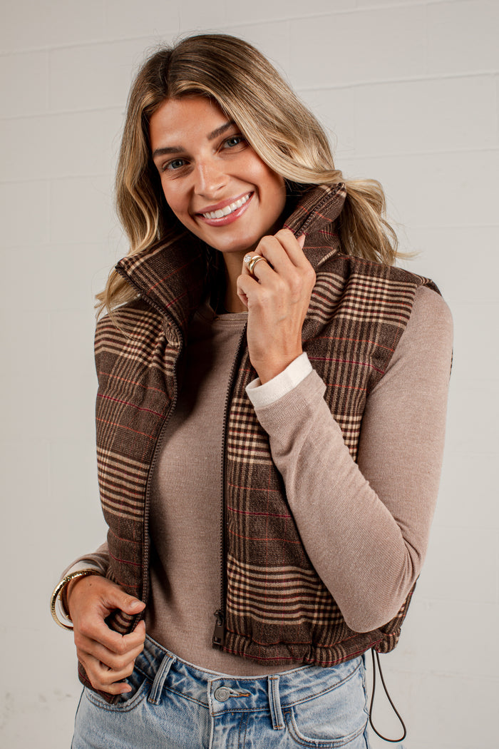 A smiling woman wearing a brown and beige plaid sleeveless puffer vest over a taupe long-sleeve top and blue jeans, standing against a light gray paneled wall.