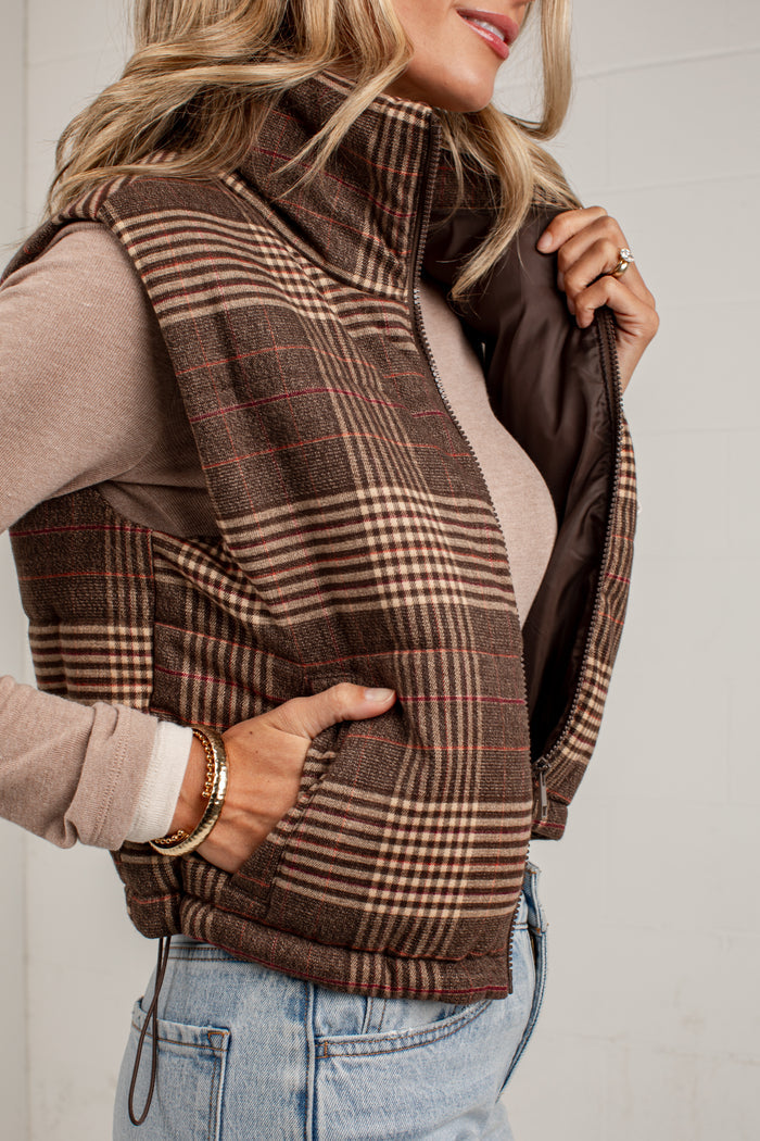 A woman wearing a brown plaid zip-front vest over a beige long-sleeve top and light blue jeans, shown in a close-up lifestyle shot against a white wall.