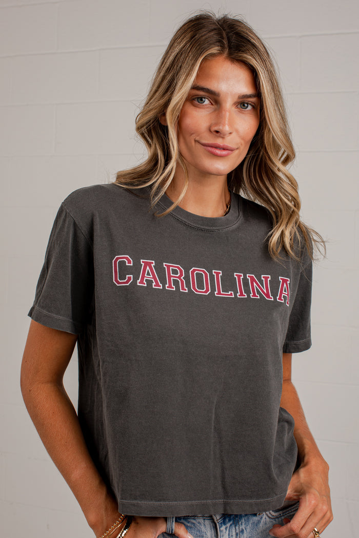 A woman wearing a dark gray washed t-shirt with 'CAROLINA' printed across the chest in red and white block letters, paired with blue jeans, standing against a white brick wall.
