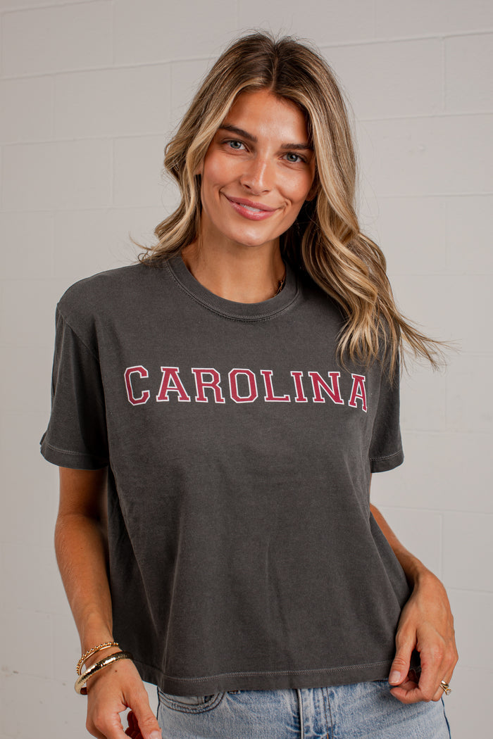 A woman wearing a dark charcoal gray short-sleeve t-shirt with 'CAROLINA' printed across the chest in red varsity-style letters with a white outline, paired with light blue jeans.