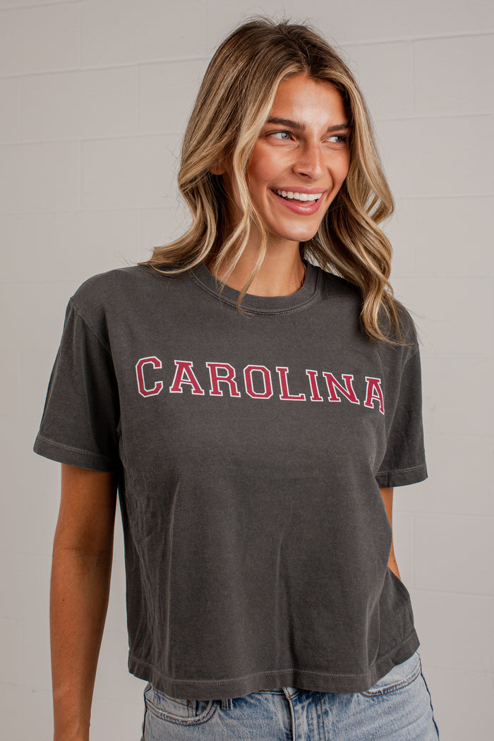 A woman wearing a dark gray washed t-shirt with 'CAROLINA' printed in red across the chest, paired with light blue jeans, standing against a light gray brick wall.