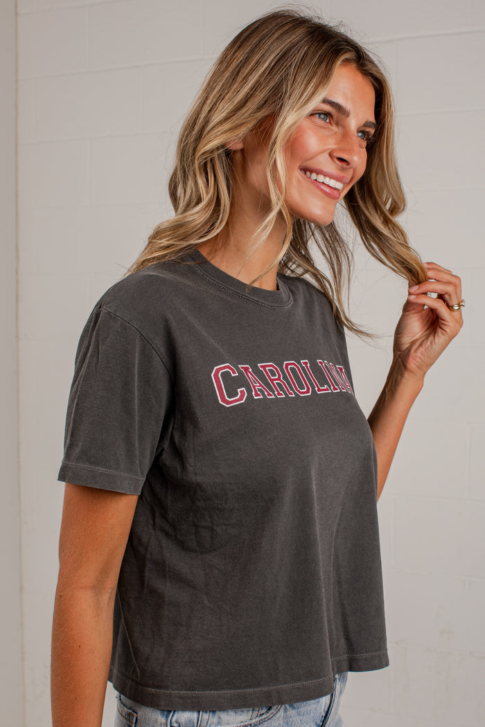 A woman wearing a dark gray washed short-sleeve t-shirt with 'CAROLINA' printed in red across the chest, paired with light blue jeans, standing against a white brick wall.