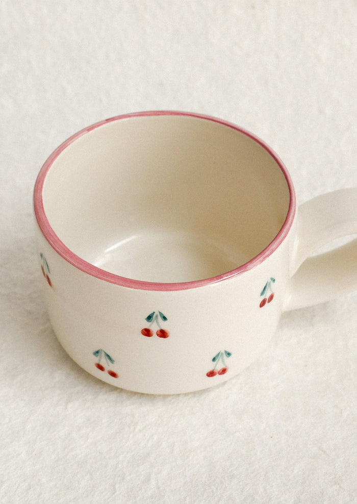 A cream ceramic mug with hand painted cherry motif and pink top rim.