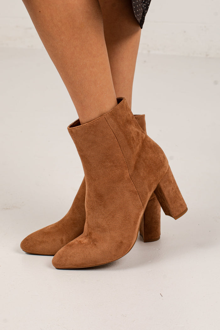 A pair of women's ankle boots in a warm camel brown suede, shown on a model's feet against a light gray studio background.