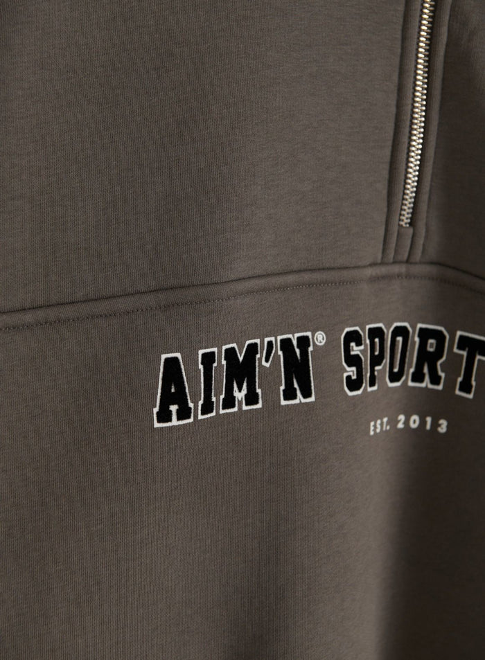 Lähikuva tummanvihreästä Varsity Sweat Half Zip -collegepuserosta, jossa näkyvät vetoketju, pehmeä kangas ja aim'n-varsity-kirjonta rinnassa
