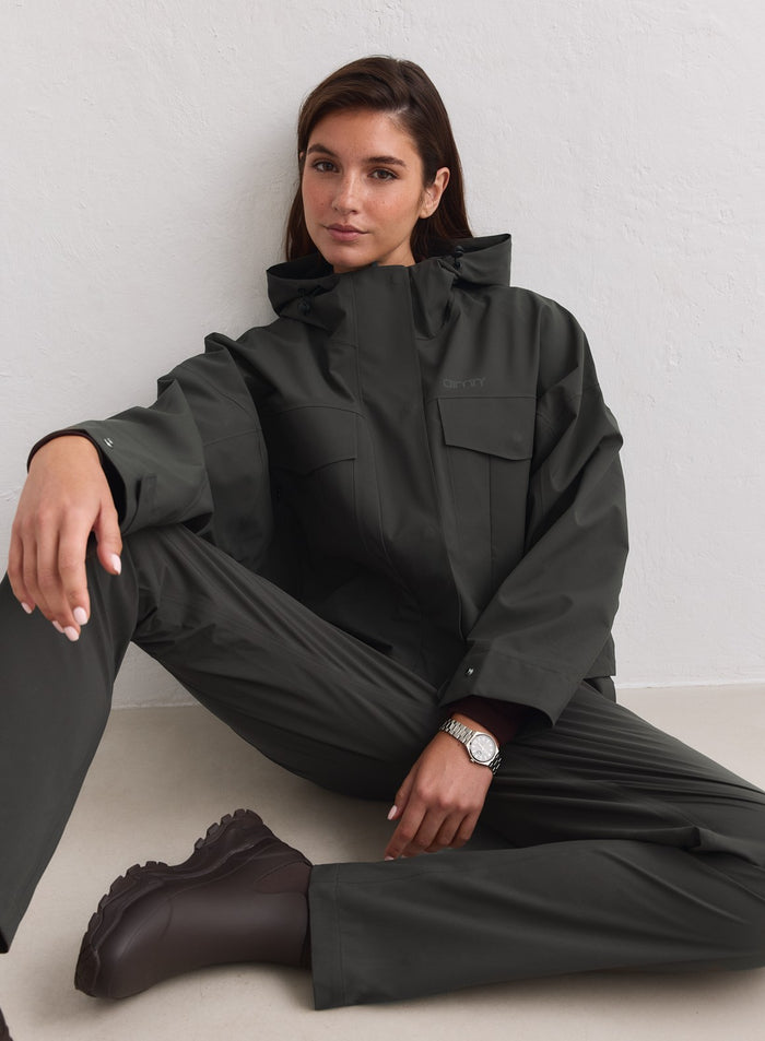 Tummanvihreä aim'n Waterproof Jacket cargo-taskuilla lattialla istuvan mallin yllä. [M-178cm]