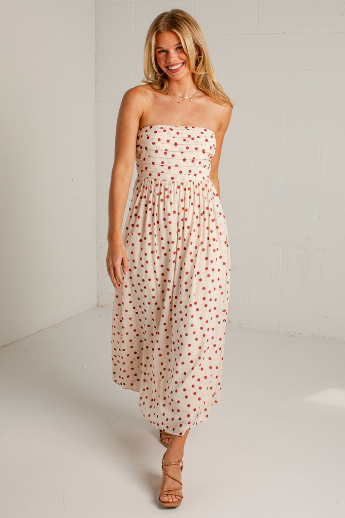 Women's cream strapless midi dress with red polka dots, a fitted bodice and a flowing skirt, styled with strappy heels.