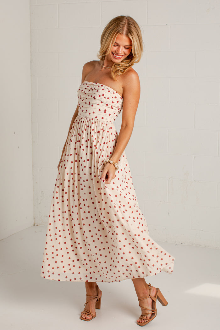 Women's cream strapless midi dress with red polka dots, a fitted bodice and a flowing skirt, styled with strappy heels.