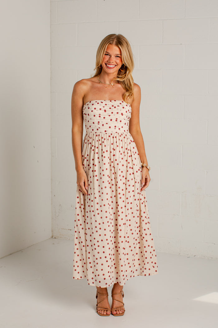 Women's cream strapless midi dress with red polka dots, a fitted bodice and a flowing skirt, styled with strappy heels.