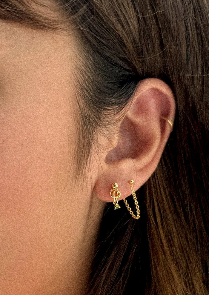 A person wearing a pair of ball stud earrings with knot and green crystal.
