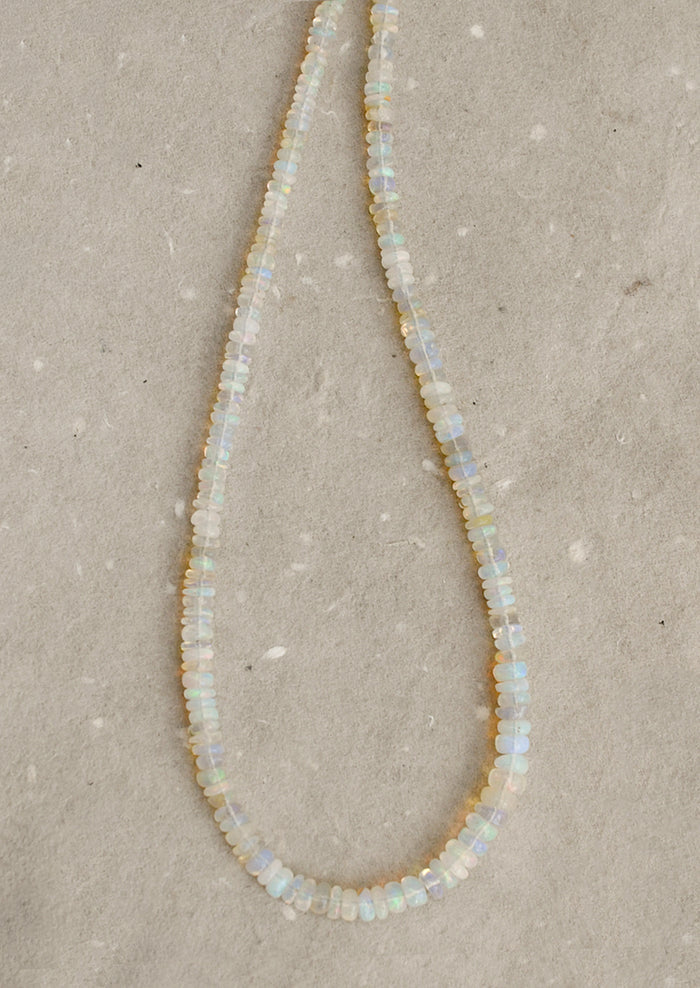 A beaded necklace made of small, irregular opal-like beads that shimmer with iridescent colors of white, pale blue, and soft yellow, laid out on a textured beige surface.