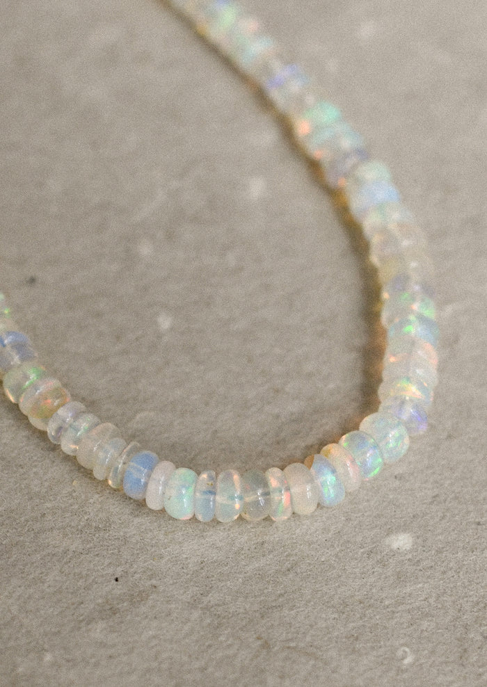 A beaded necklace made of small, irregular opal-like beads that shimmer with iridescent colors of white, pale blue, and soft yellow, laid out on a textured beige surface.