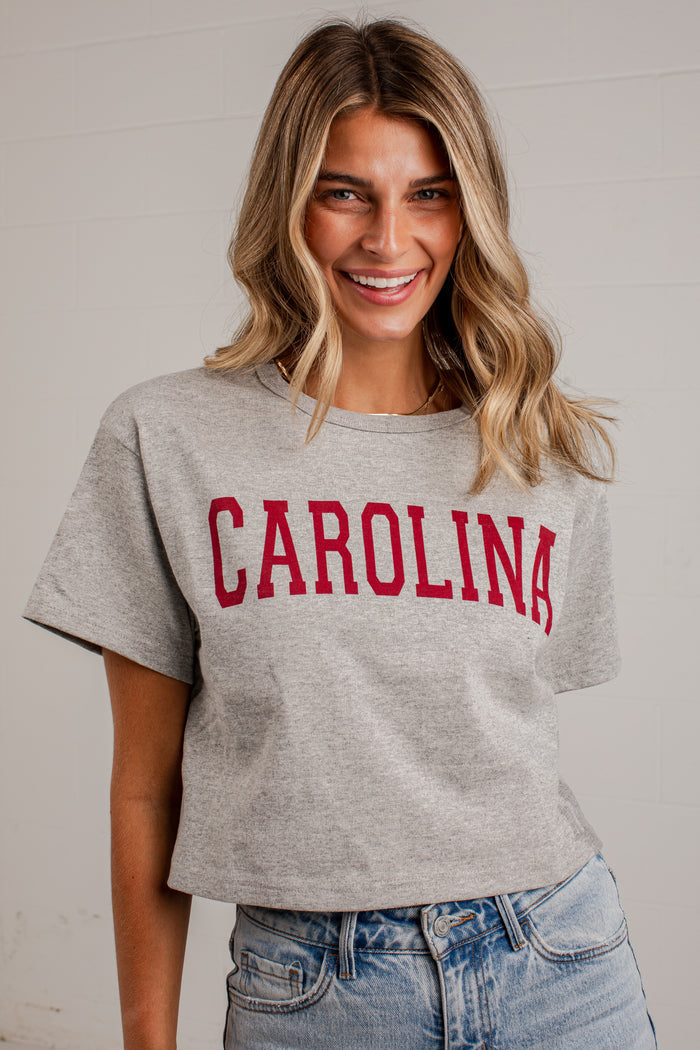 A woman wearing a heather gray cropped t-shirt with 'CAROLINA' printed in red arched block letters across the chest, paired with light wash blue jeans.
