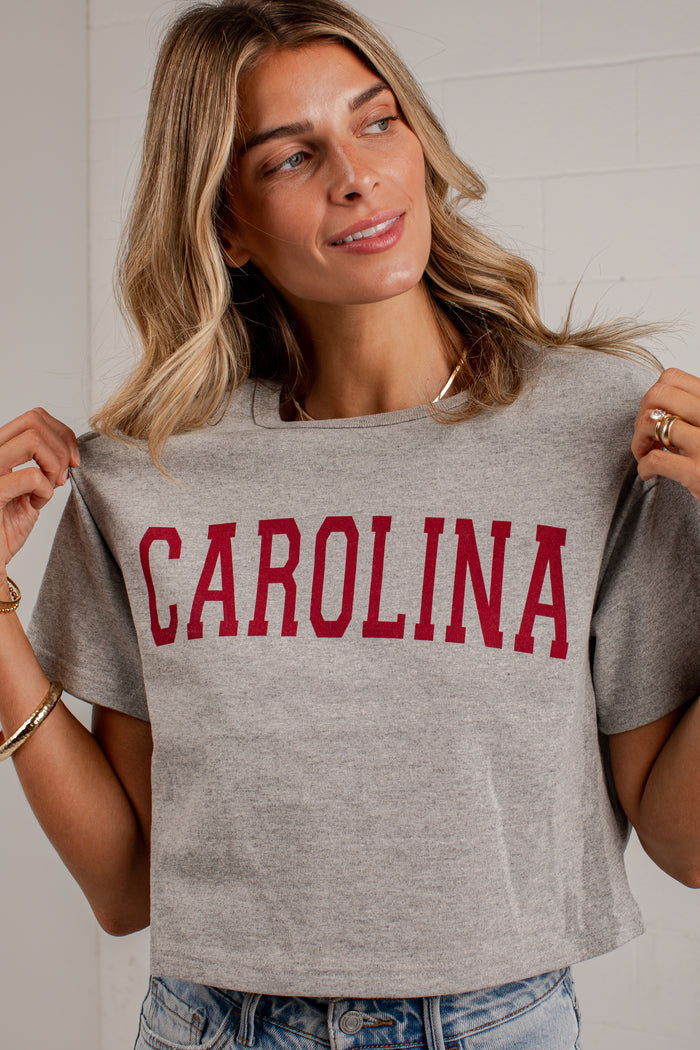 A woman wearing a heather gray cropped t-shirt with 'CAROLINA' printed in red across the chest, holding the shirt outwards to show its fit. She is wearing blue jeans and gold jewelry, standing against a white brick wall.
