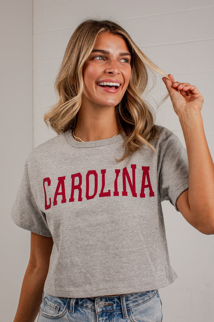 A woman wearing a heather gray cropped t-shirt with 'CAROLINA' printed in red across the chest, paired with blue jeans, standing against a white paneled wall.