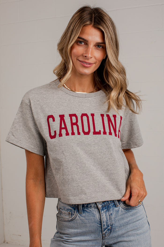 A woman wearing a heather gray cropped t-shirt with 'CAROLINA' printed in red arched letters across the chest, paired with light-wash blue jeans.