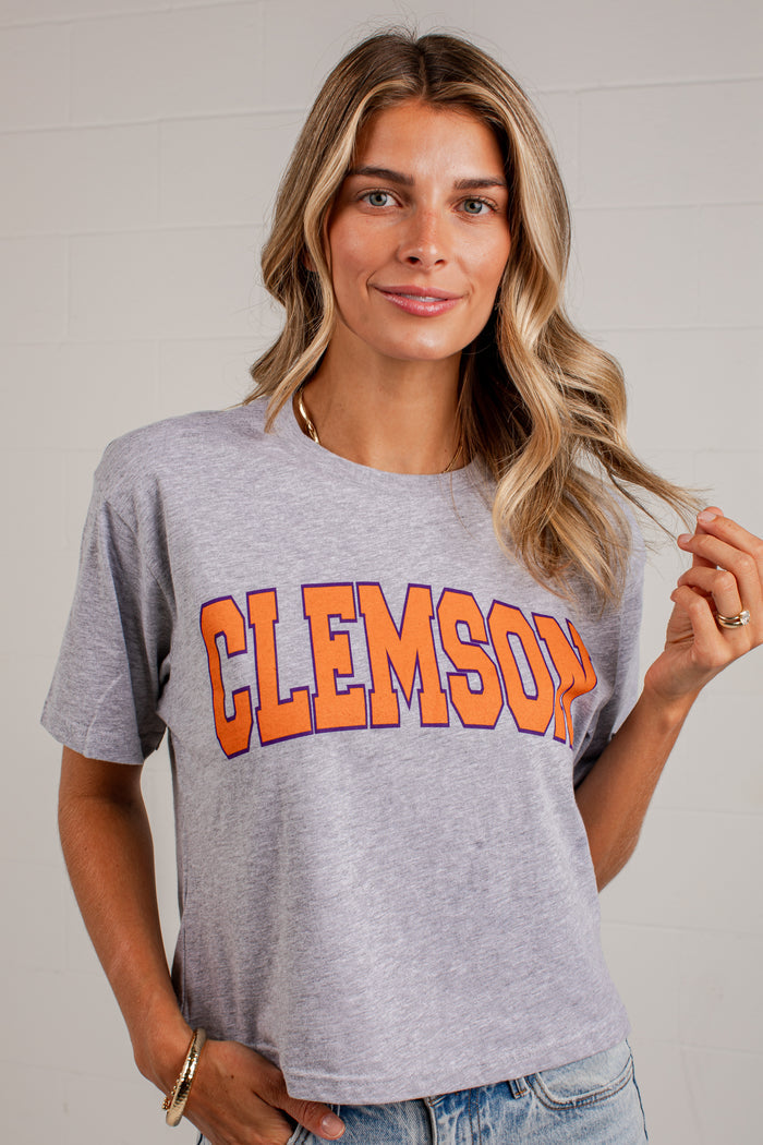 A woman wearing a heather gray Clemson Tigers graphic t-shirt with 'CLEMSON' printed in orange and purple varsity letters, paired with blue jeans against a white paneled wall.