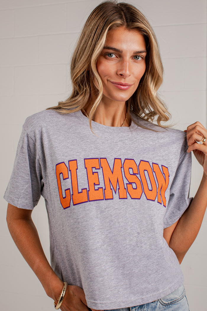 A woman wearing a heather gray Clemson athletic t-shirt with 'CLEMSON' printed in orange and blue varsity block letters across the chest, holding the fabric outwards to show its drape.