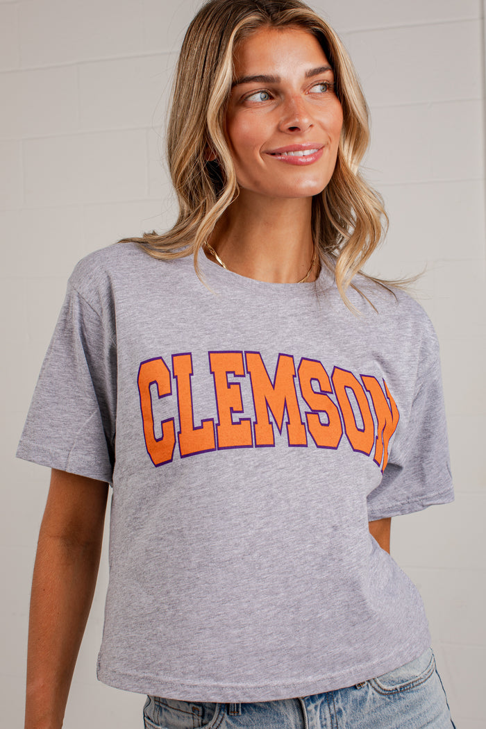 A woman wearing a heather gray short-sleeve t-shirt with 'CLEMSON' printed in orange and navy blue varsity lettering across the chest, paired with light-wash blue jeans.