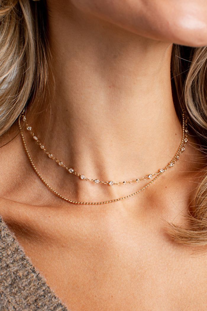 A close-up of a model's neck and collarbone wearing two delicate rose gold chain necklaces, one with small clear stones and one with a fine beaded chain.