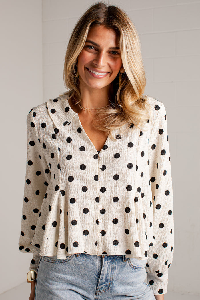 Women's cream blouse with black polka dots, a V-neckline, front buttons and long sleeves, styled with light-wash jeans.