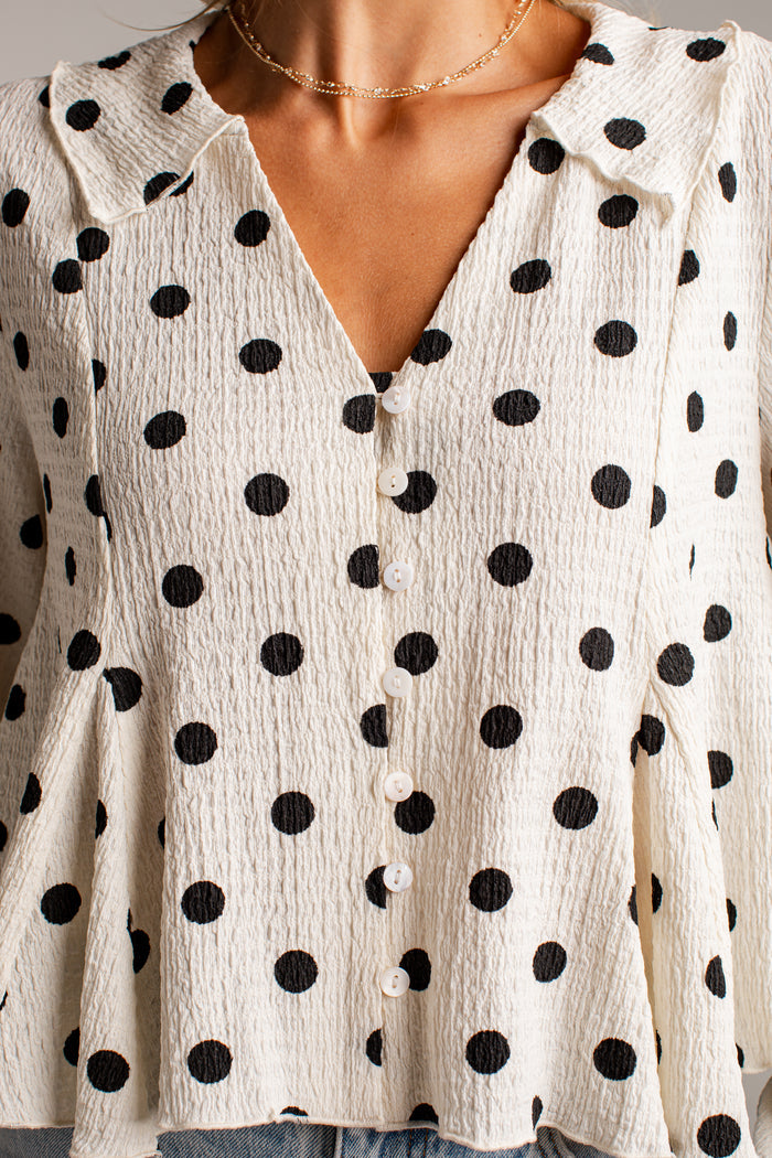 Women's cream blouse with black polka dots, a V-neckline, front buttons and long sleeves, styled with light-wash jeans.