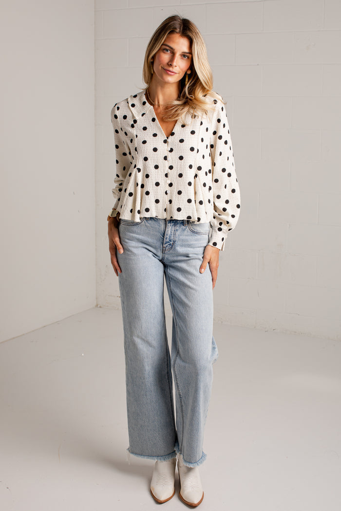 Women's cream blouse with black polka dots, a V-neckline, front buttons and long sleeves, styled with light-wash jeans.