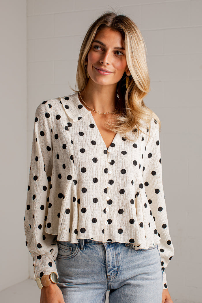 Women's cream blouse with black polka dots, a V-neckline, front buttons and long sleeves, styled with light-wash jeans.