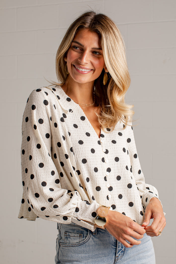 Women's cream blouse with black polka dots, a V-neckline, front buttons and long sleeves, styled with light-wash jeans.