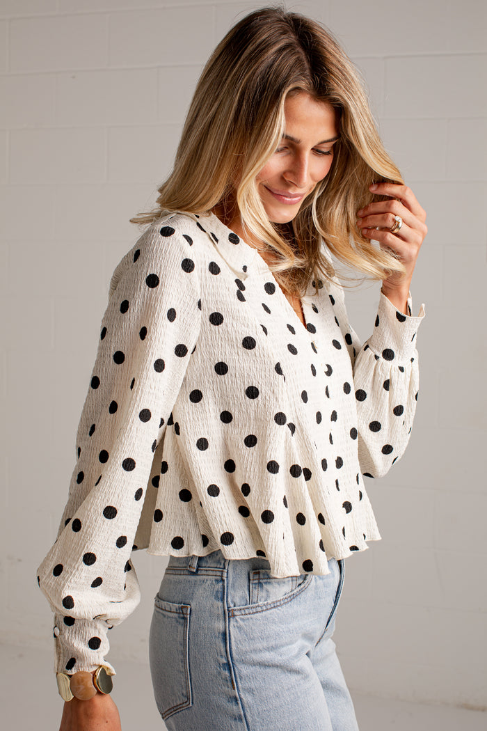 Women's cream blouse with black polka dots, a V-neckline, front buttons and long sleeves, styled with light-wash jeans.