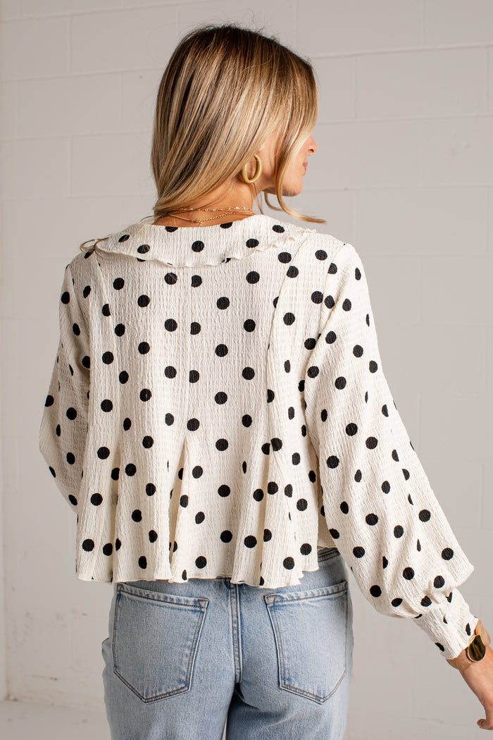 Women's cream blouse with black polka dots, a V-neckline, front buttons and long sleeves, styled with light-wash jeans.