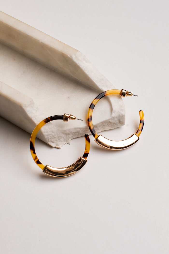 Tortoiseshell hoop earrings with gold-toned metal accents, displayed on a light gray surface next to a white stone prop.