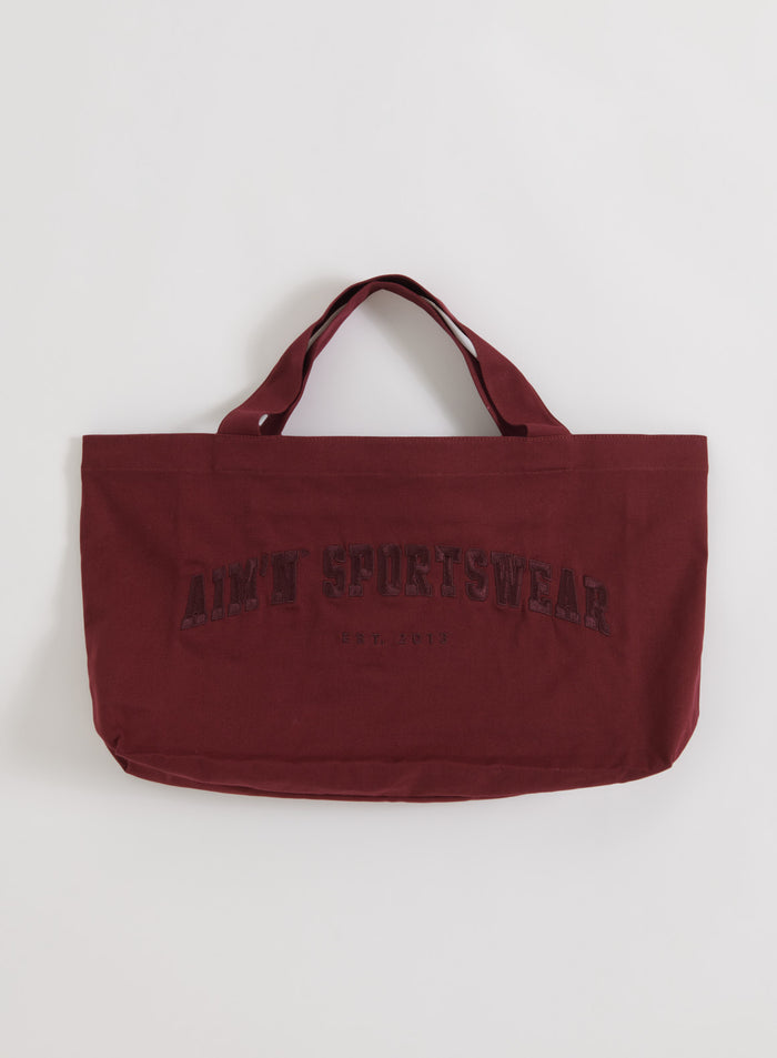 Puuvillainen Merlot Varsity Tote Bag sävyyn sopivalla aim'n sportswear -logolla tasokuvassa.