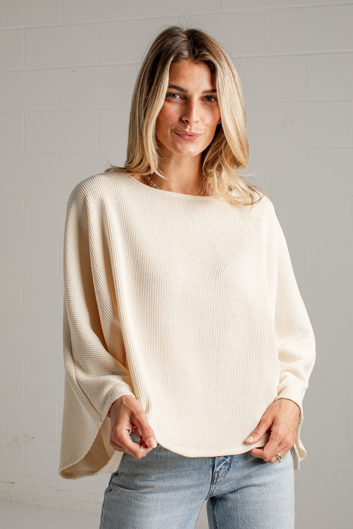A woman wearing a cream-colored, long-sleeved ribbed knit top with a wide boat neckline and draped sleeves, paired with light-wash blue jeans, standing against a white brick wall.