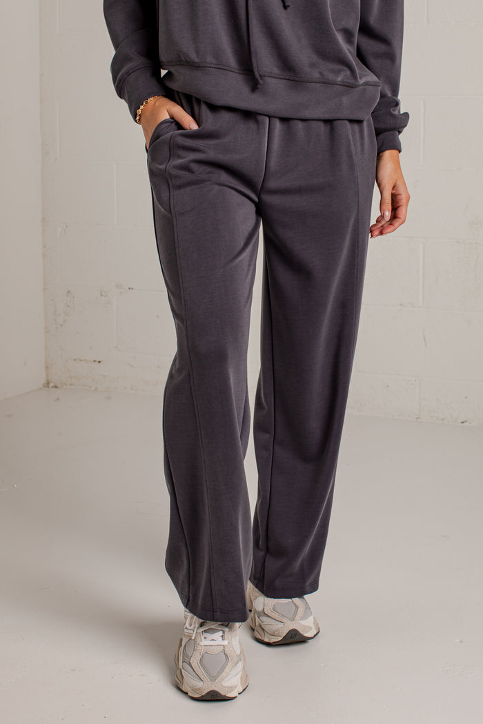 A person wearing dark charcoal gray sweatpants with a matching sweatshirt and white sneakers, standing in a minimalist studio setting with a white brick wall background.