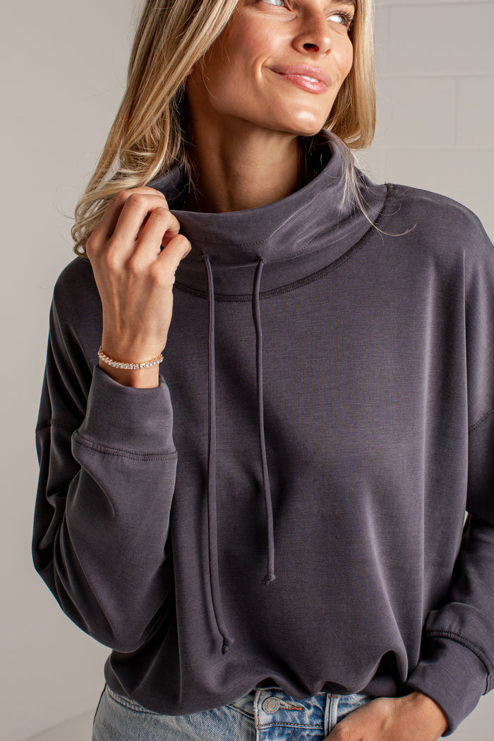 A woman wearing a dark charcoal gray cowl neck sweatshirt with drawstrings, paired with light wash blue jeans, shown in a close-up shot against a neutral light gray background.