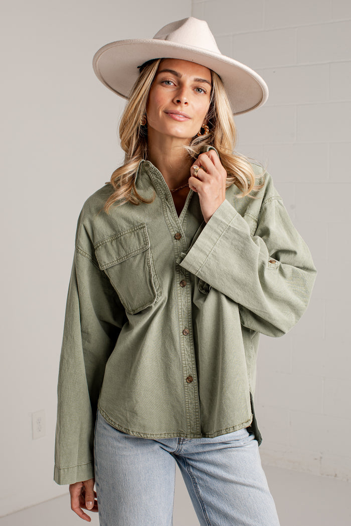 A woman wearing an oversized sage green button-up shirt with chest pockets, light-wash jeans, and a wide-brimmed cream hat, standing against a light gray wall.