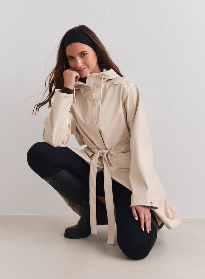 Hiekanvärinen aim'n Rain Jacket säädettävällä hupulla ja vyöllä yhdistettynä mustiin leggingseihin. [M-178cm]