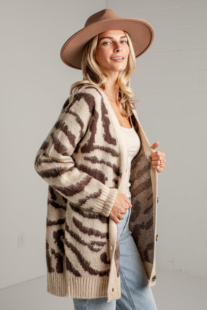 A woman wearing a beige and brown zebra-print knit cardigan, a brown wide-brim hat, a white top, and light blue jeans, standing against a light gray brick wall.