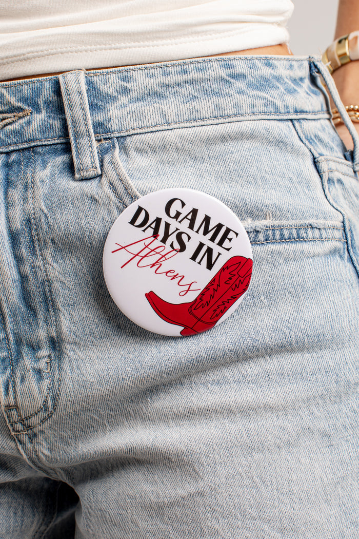 A round pin-back button badge with the text 'GAME DAYS IN Athens' and a red boot graphic, pinned to the back pocket of light-wash blue jeans.