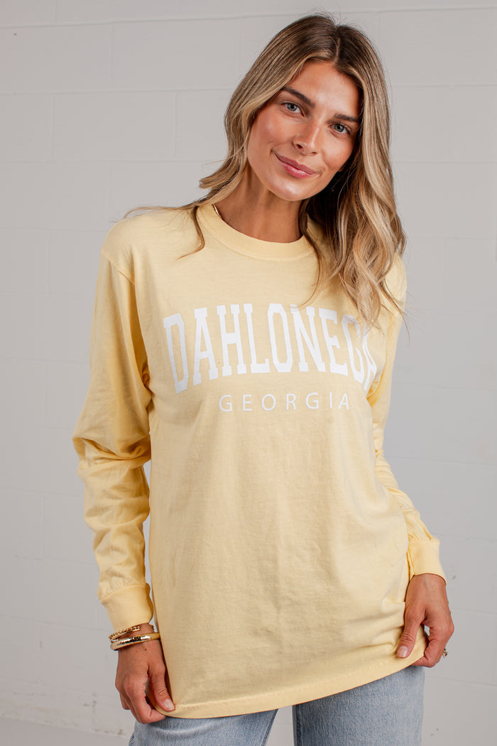 A woman wearing a pale yellow long-sleeve t-shirt with 'DAHLONEGA GEORGIA' printed in white across the chest, paired with light blue jeans, standing against a white brick wall.